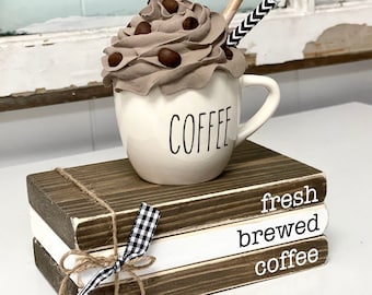 May include: A white ceramic mug with the word "COFFEE" printed on it, filled with faux whipped cream and chocolate sprinkles, topped with a brown and white striped straw. The mug is sitting on top of three stacked wooden blocks with the words "fresh brewed coffee" painted on them in white.