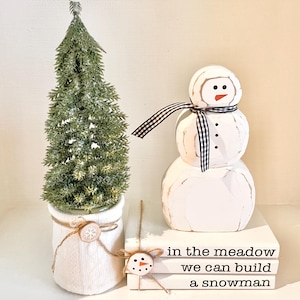 May include: A decorative winter scene featuring a small green Christmas tree in a white pot, a wooden snowman with a black and white scarf, and a stack of books with the text "in the meadow we can build a snowman."