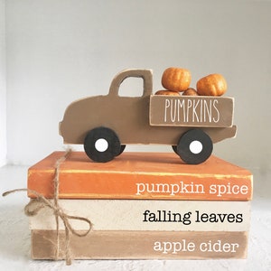 May include: A brown wooden toy truck with white wheels is carrying a stack of orange pumpkins. The truck is sitting on top of three stacked wooden blocks. The blocks are painted in shades of orange, beige, and brown. The top block has the text "pumpkin spice" written on it. The middle block has the text "falling leaves" written on it. The bottom block has the text "apple cider" written on it. The truck has the word "PUMPKINS" written on the side.