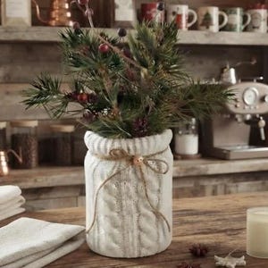 May include: A white ceramic jar with a cable-knit texture, wrapped in twine with a small wooden bead, holding a bouquet of green pine branches and dark red berries. The jar sits on a wooden surface with small white crocheted ornaments.