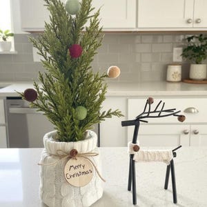 May include: A small evergreen tree in a white cable-knit sweater pot with a round wooden tag reading Merry Christmas, decorated with felt balls, alongside a black metal reindeer figurine wearing a matching white cable-knit sweater piece.