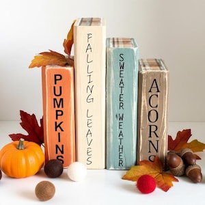 May include: Four rectangular wooden blocks arranged in a row, painted orange, cream, blue, and tan. The blocks feature the black text PUMPKINS, FALLING LEAVES, SWEATER WEATHER, and ACORNS. They are decorated with plaid tops, felt balls, and faux leaves.