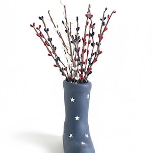 May include: A navy blue boot-shaped vase with white star accents holds a bundle of decorative branches. The branches feature small red, white, and navy blue berries, creating a patriotic theme. The vase is a decorative home accent.