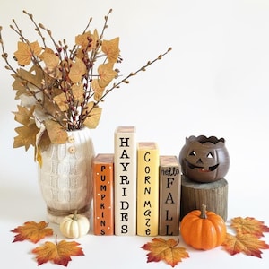 May include: A white cable-knit vase filled with autumn branches, four rectangular wooden blocks labeled PUMPKINS, HAYRIDES, CORN MAZE, and HELLO FALL, a brown jack-o'-lantern candle holder on a log, and small orange and white pumpkins.