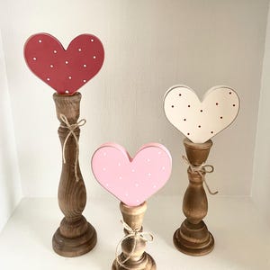 Pode incluir: Tr&ecirc;s decora&ccedil;&otilde;es de cora&ccedil;&atilde;o de madeira em vermelho, rosa e creme, cada uma com bolinhas brancas ou vermelhas. Os cora&ccedil;&otilde;es s&atilde;o montados em casti&ccedil;ais de madeira torneada, amarrados com barbante. Um charmoso item de decora&ccedil;&atilde;o r&uacute;stica.