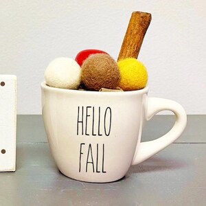 Pode incluir: Caneca de cer&acirc;mica branca com o texto "Hello Fall" em letras pretas. A caneca est&aacute; cheia de bolas de feltro marrom, branco e amarelo e um pau de canela.