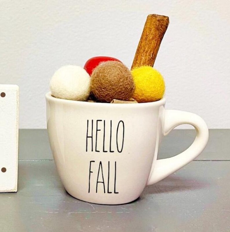 Pode incluir: Caneca de cer&acirc;mica branca com o texto "Hello Fall" em letras pretas. A caneca est&aacute; cheia de bolas de feltro marrom, branco e amarelo e um pau de canela.