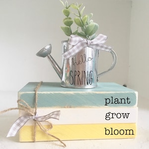 May include: Three stacked wooden blocks painted in yellow, white, and blue with the words "plant", "grow", and "bloom" written on them. A small metal watering can with the words "hello spring" is on top of the blocks.