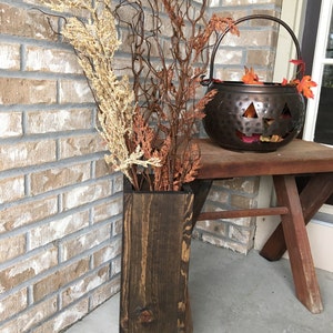 May include: A dark brown wooden vase with a rustic finish, filled with dried branches and flowers in shades of beige and orange. The vase is standing on a wooden porch with a brick wall in the background.