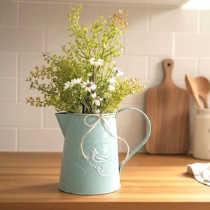 May include: A light blue metal pitcher with a handle, filled with greenery and white flowers. The pitcher has a bird and text design and is tied with twine. The pitcher is on a wooden surface.