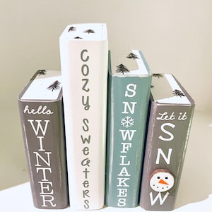 May include: Four decorative wooden blocks painted in winter colors. The blocks read "hello WINTER", "Cozy Sweaters", "SNOWFLAKES", and "Let it SNOW". Each block has a different design, including a snowman and pine trees.