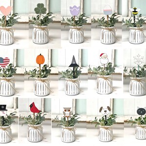May include: A set of 16 decorative plant picks featuring various designs, including hearts, pumpkins, snowflakes, and animals. The picks are inserted into small white pots with a rustic striped design.