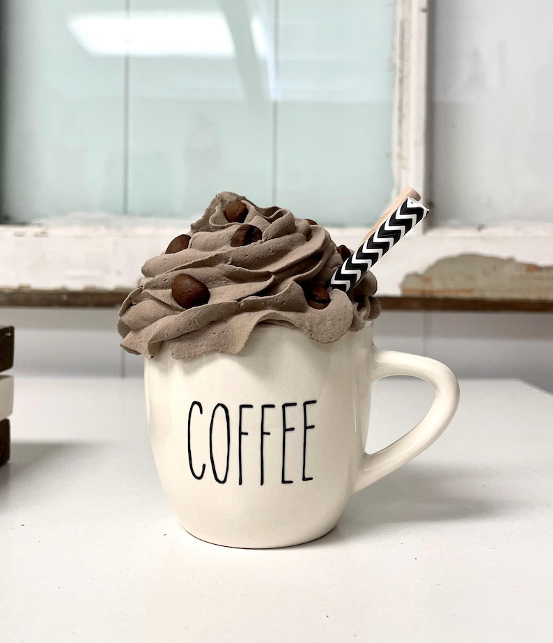 May include: A white ceramic coffee mug with the word "COFFEE" printed on the front. The mug is filled with a brown whipped cream topping with chocolate sprinkles and a black and white striped straw.