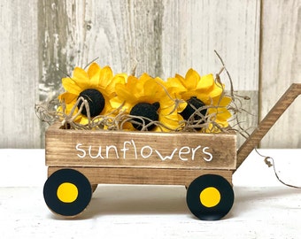 Könnte beinhalten: Ein Holzkarren mit schwarzen Rädern und gelben Akzenten, gefüllt mit drei gelben Filz-Sonnenblumen. Der Karren hat das Wort "Sunflowers" an der Seite bemalt.