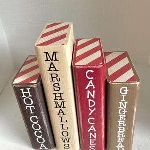 K&ouml;nnte beinhalten: Vier Holzbl&ouml;cke in verschiedenen Farben, die jeweils einem Buch &auml;hneln. Die Bl&ouml;cke sind mit den W&ouml;rtern "Hot Cocoa", "Marshmallows", "Candy Canes" und "Gingerbread" in wei&szlig;er Schrift beschriftet. Die Oberseiten weisen ein rot-wei&szlig; gestreiftes Muster auf.