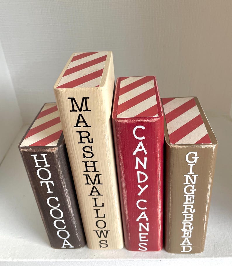 K&ouml;nnte beinhalten: Vier Holzbl&ouml;cke in verschiedenen Farben, die jeweils einem Buch &auml;hneln. Die Bl&ouml;cke sind mit den W&ouml;rtern "Hot Cocoa", "Marshmallows", "Candy Canes" und "Gingerbread" in wei&szlig;er Schrift beschriftet. Die Oberseiten weisen ein rot-wei&szlig; gestreiftes Muster auf.