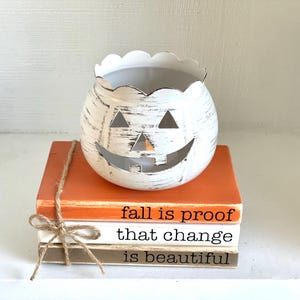 May include: A white, distressed pumpkin-shaped candle holder with a carved face sits atop a stack of decorative books. The books are orange, white, and brown, tied with twine, and feature the text "fall is proof that change is beautiful."