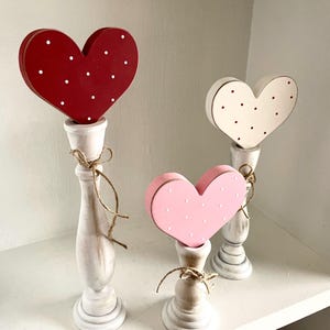 May include: Three wooden heart decorations in red, pink, and white, each with white polka dots. The hearts are mounted on white candlestick holders and tied with twine. A charming, decorative accent for Valentine's Day or other special occasions.