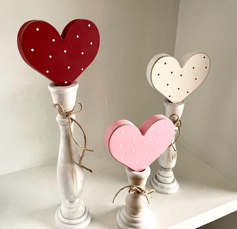 May include: Three wooden heart decorations in red, pink, and white, each with white polka dots. The hearts are mounted on white candlestick holders and tied with twine. A charming, decorative accent for Valentine's Day or other special occasions.