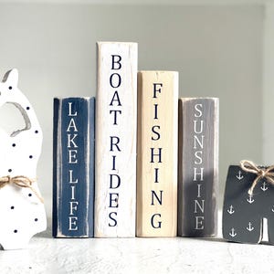 May include: A collection of wooden beach-themed decorations. Includes a white swimsuit shape with black polka dots, wooden blocks with the words "LAKE LIFE", "BOAT RIDES", "FISHING", and "SUNSHINE", and a pair of gray shorts with white anchor designs.