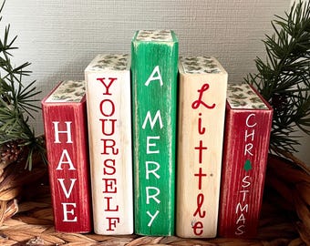 Könnte beinhalten: Fünf Holzblöcke, rot, weiß und grün bemalt, angeordnet, um "Have Yourself a Merry Little Christmas" zu buchstabieren. Die Blöcke haben ein antikes Finish und sind mit festlichen Mustern und Texten verziert. Ein Weihnachtsbaum und Tannennadeln sind sichtbar.