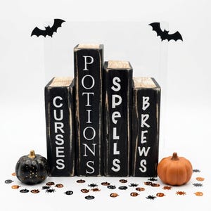 May include: Four black wooden blocks shaped like book spines with distressed edges, featuring white text that reads CURSES, POTIONS, SPELLS, and BREWS. The set includes two black bat silhouettes, a black glittery pumpkin, and an orange pumpkin.