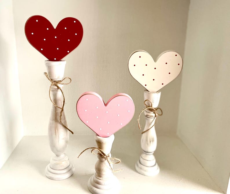 May include: Three decorative heart-shaped candle holders. The hearts are red, pink, and off-white, each with white or black polka dots. The candle holders are white with a distressed finish and tied with twine.