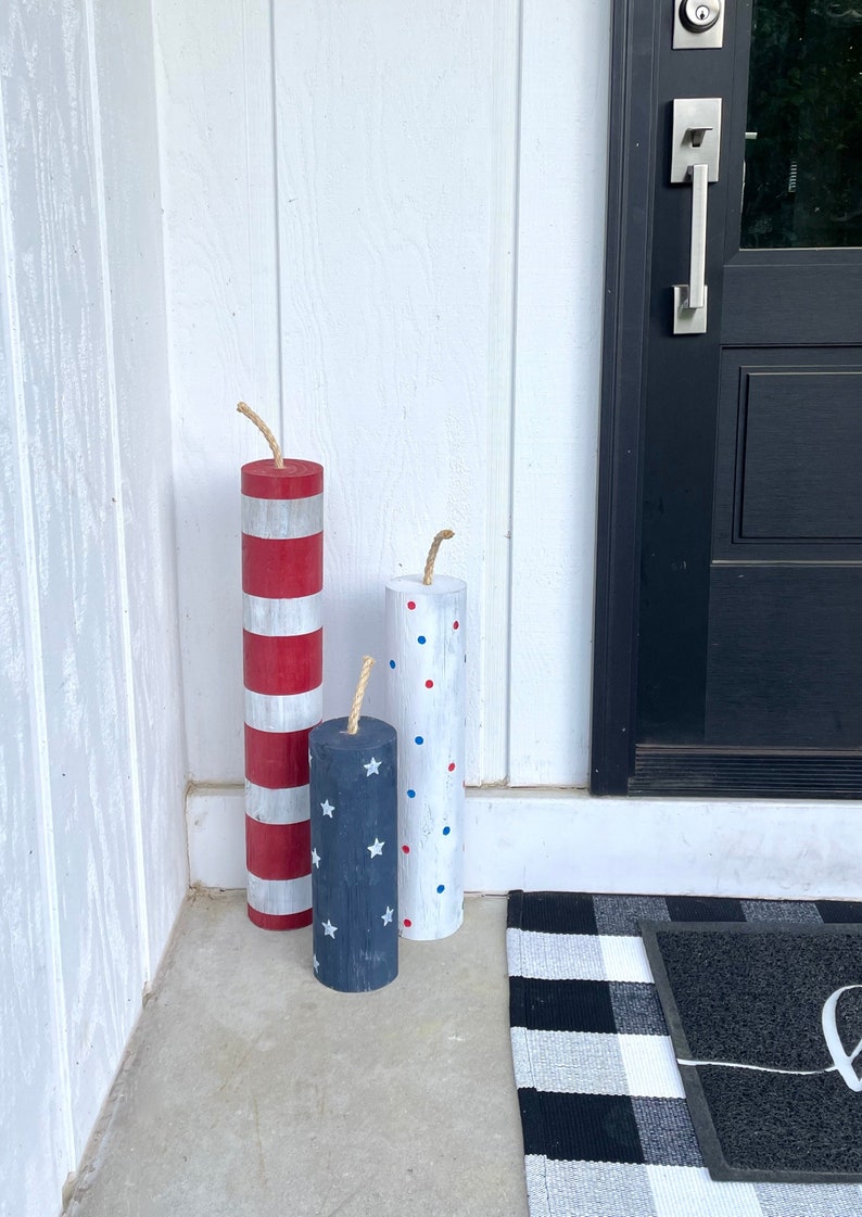 May include: Three wooden firework decorations painted in red, white, and blue with stars and polka dots. The decorations are placed on a concrete porch with a black and white checkered doormat.