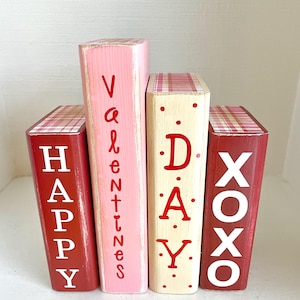 May include: Four wooden blocks painted in red, pink, and cream colors. The blocks spell out "Happy Valentines Day XOXO" in white and red lettering. The tops of the blocks have a red and white plaid pattern.
