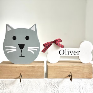May include: A set of two wooden Christmas stocking holders. One is shaped like a gray cat with white whiskers and black eyes. The other is shaped like a white bone with a red and black plaid bow and the name "Oliver" written on it.
