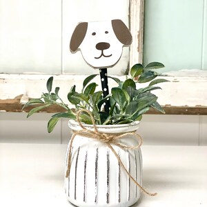 May include: White ceramic planter with vertical lines and a brown and white wooden dog head sticking out of the top. The planter is tied with twine and filled with green leafy plants.
