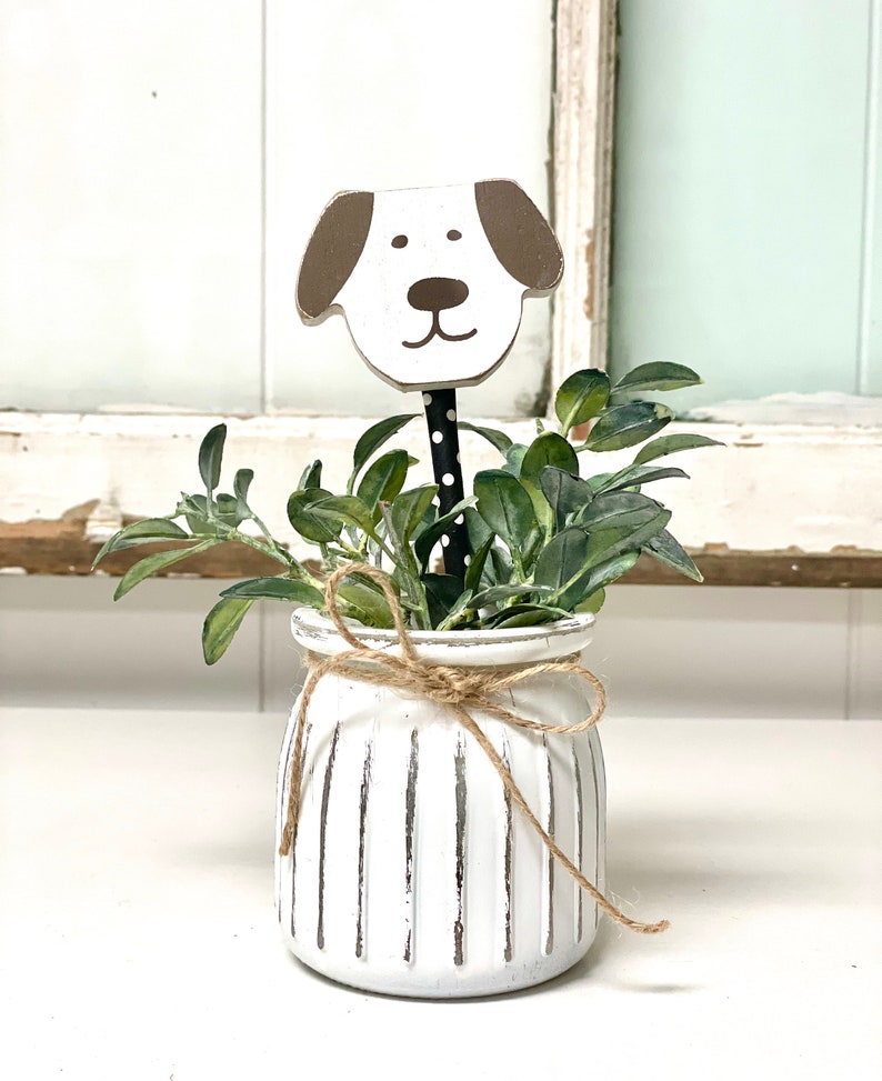 May include: White ceramic planter with vertical lines and a brown and white wooden dog head sticking out of the top. The planter is tied with twine and filled with green leafy plants.