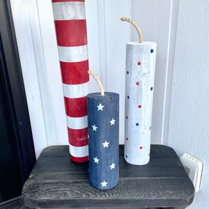 May include: Three wooden firework decorations with red, white, and blue paint. The decorations are striped, polka dotted, and have a star pattern. They are sitting on a black wooden table.
