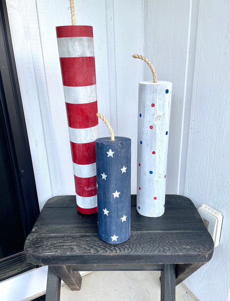 May include: Three wooden firework decorations with red, white, and blue paint. The decorations are striped, polka dotted, and have a star pattern. They are sitting on a black wooden table.