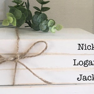 May include: Three white books stacked on top of each other. The books are tied together with twine and have the names "Nick", "Logan", and "Jack" written on them.