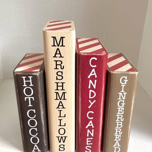 K&ouml;nnte beinhalten: Vier Holzbl&ouml;cke, die wie B&uuml;cher bemalt sind. Jeder Block hat eine andere Farbe: braun, Naturholz, rot und beige. Die Bl&ouml;cke haben wei&szlig;en Text mit der Aufschrift "Hot Cocoa", "Marshmallows", "Candy Canes" und "Gingerbread". Die Oberseiten haben rot-wei&szlig;e Streifen.