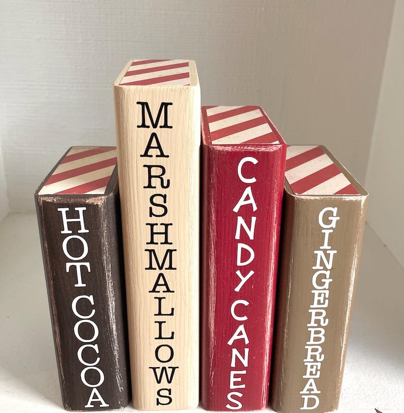 K&ouml;nnte beinhalten: Vier Holzbl&ouml;cke, die wie B&uuml;cher bemalt sind. Jeder Block hat eine andere Farbe: braun, Naturholz, rot und beige. Die Bl&ouml;cke haben wei&szlig;en Text mit der Aufschrift "Hot Cocoa", "Marshmallows", "Candy Canes" und "Gingerbread". Die Oberseiten haben rot-wei&szlig;e Streifen.