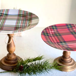 May include: Two decorative cake stands with plaid patterned plates. One plate has a red, green, and white pattern, and the other has a red, green, and yellow pattern. The stands have wooden bases and are decorated with greenery.