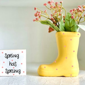 May include: A white wooden sign with pink polka dots reads "Spring has sprung". A yellow rain boot with pink polka dots is filled with pink flowers.
