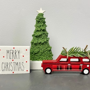 May include: A red wooden car with a plaid design is carrying a small green Christmas tree on top. The car is next to a white wooden sign that says "Merry Christmas" in black lettering with red dots.
