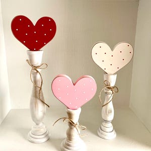 May include: Three decorative heart-shaped objects on white candle holders. The hearts are red, pink, and white, each with white or red polka dots. The candle holders are white with a distressed finish, and tied with twine.