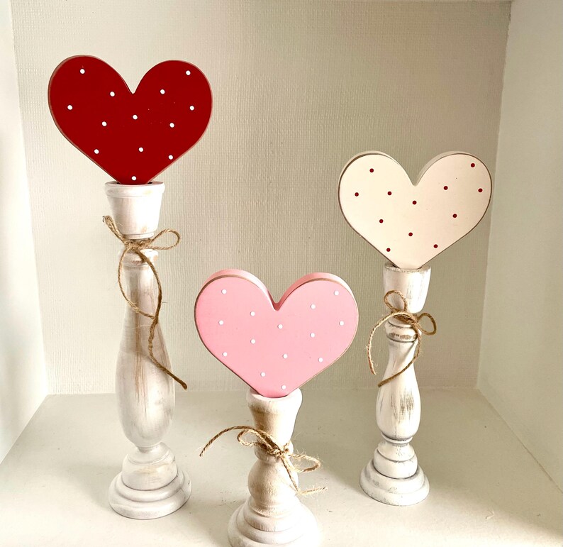 May include: Three decorative heart-shaped objects on white candle holders. The hearts are red, pink, and white, each with white or red polka dots. The candle holders are white with a distressed finish, and tied with twine.