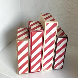 K&ouml;nnte beinhalten: Eine Ansammlung von f&uuml;nf rechteckigen Schachteln mit rot-wei&szlig;en diagonalen Streifen. Die unterschiedlich gro&szlig;en Schachteln sind versetzt angeordnet. Die Schachteln haben eine Vintage-&Auml;sthetik.