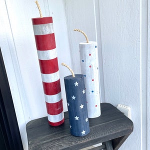 May include: Three wooden, cylindrical, firework-shaped decorations painted in red, white, and blue patriotic colors. The decorations are sitting on a black wooden table. The decorations are striped, polka dotted, and have white stars on a blue background.