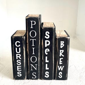 May include: Four black wooden blocks with white lettering spelling "Curses", "Potions", "Spells", and "Brews". The tops of the blocks have a light brown, wood-grain appearance. The blocks are arranged in a staggered row.