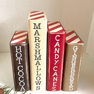 K&ouml;nnte beinhalten: Vier Holzbl&ouml;cke, die wie B&uuml;cher bemalt sind, mit den W&ouml;rtern "Hot Cocoa", "Marshmallows", "Candy Canes" und "Gingerbread" in wei&szlig;en Buchstaben. Jeder Block hat ein rot-wei&szlig; gestreiftes Muster auf der Oberseite.