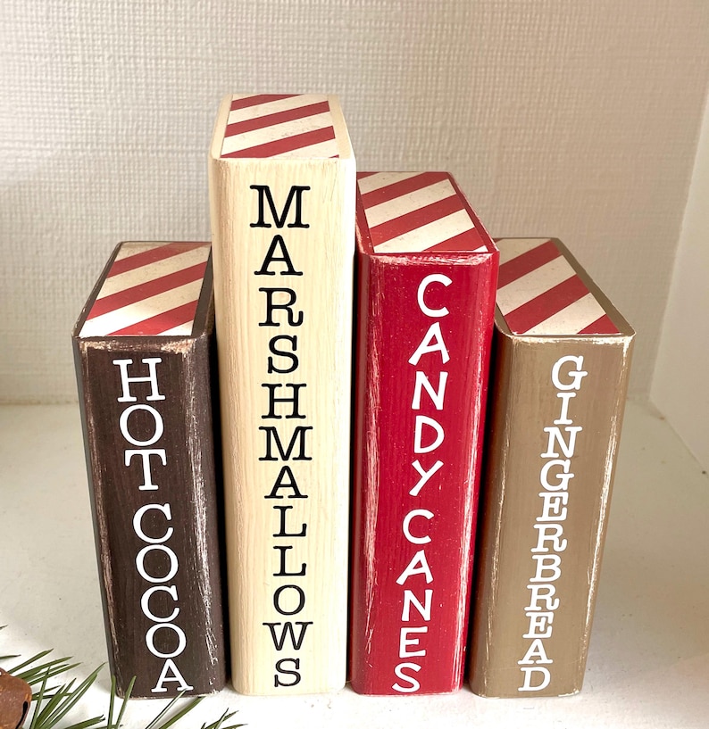 K&ouml;nnte beinhalten: Vier Holzbl&ouml;cke, die wie B&uuml;cher bemalt sind, mit den W&ouml;rtern "Hot Cocoa", "Marshmallows", "Candy Canes" und "Gingerbread" in wei&szlig;en Buchstaben. Jeder Block hat ein rot-wei&szlig; gestreiftes Muster auf der Oberseite.