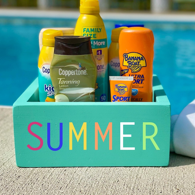 Pode incluir: Uma caixa de madeira turquesa com a palavra "SUMMER" em letras coloridas na frente. A caixa cont&eacute;m v&aacute;rios frascos de protetor solar, incluindo Coppertone Tanning Lotion, Banana Boat Sport Sunscreen e uma garrafa de protetor solar com a etiqueta "Family Size".