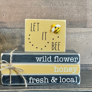 May include: A decorative wooden block with the phrase "LET IT BEE" and a 3D bee. Below are three stacked wooden blocks with the words "wild flower", "honey", and "fresh & local". The blocks are tied together with twine.