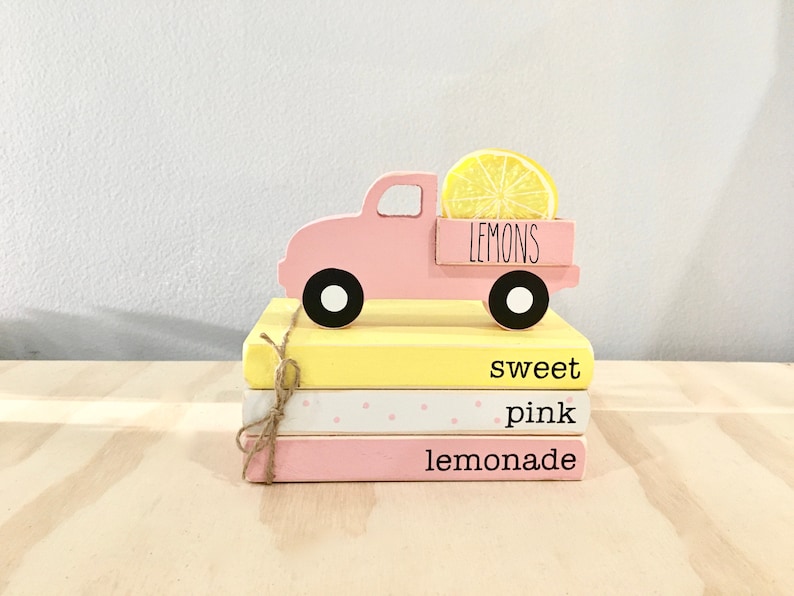 May include: A pink wooden toy truck with a yellow lemon on top. The truck is sitting on a stack of three books. The books are yellow, white with pink polka dots, and pink. The books are labeled "sweet", "pink", and "lemonade".